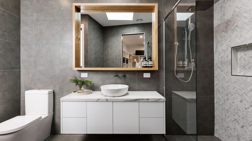 A completed bathroom renovation in Sydney with a wall-hung white vanity, marble benchtop, timber-framed mirror and grey tiled walls by 10X Joinery.
