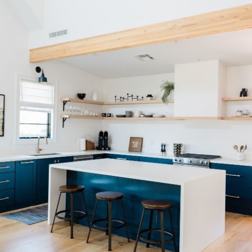 A navy blue kitchen with a white waterfall island, timber floating shelves and exposed timber beam, delivered to an agreed budget in Auburn by 10X Joinery.