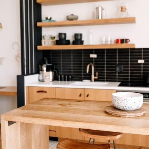 A light timber kitchen island with black tiled splashback and floating shelves, planned to connect the living areas of a Baulkham Hills home by 10X Joinery.