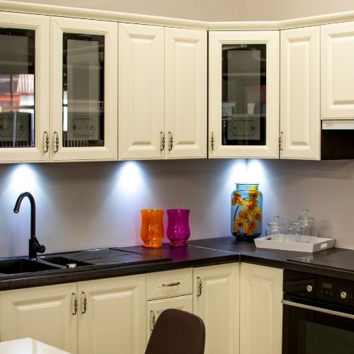 Cream kitchen cabinetry with glass-front overheads, a dark benchtop and under-cabinet lighting in an Auburn home by 10X Joinery.