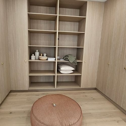 Timber walk-in robe with floor-to-ceiling open shelving and a round ottoman