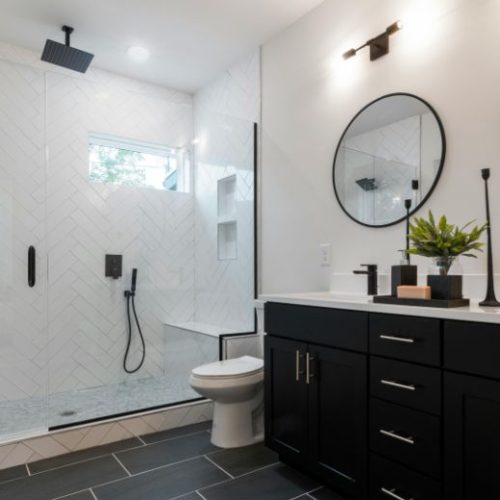 A black bathroom vanity with a round black-framed mirror and white herringbone shower tiles, designed by 10X Joinery.